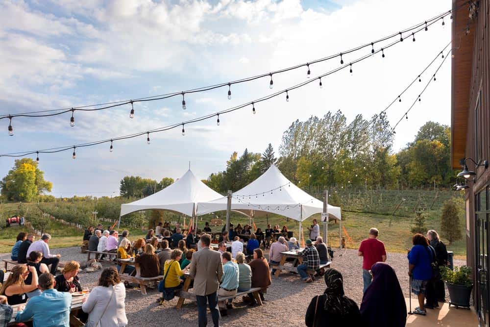 A large group of people is gathered outdoors under white tents for an event. They sit on benches and chairs, facing a speaker. String lights are hung above, and trees and greenery can be seen in the background. The sky is partly cloudy.
