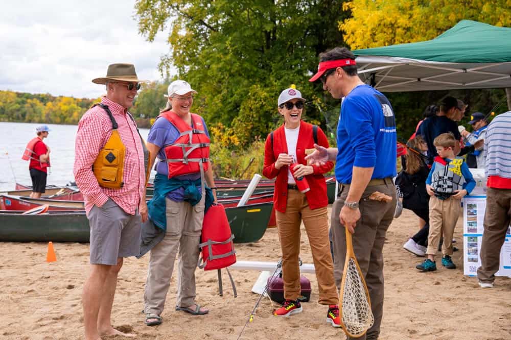 A group of five people stand on a sandy beach near a lake with canoes. They are engaged in conversation and laughter. Some wear life vests, and one person holds a fishing net. A green canopy is in the background with trees displaying fall foliage.
