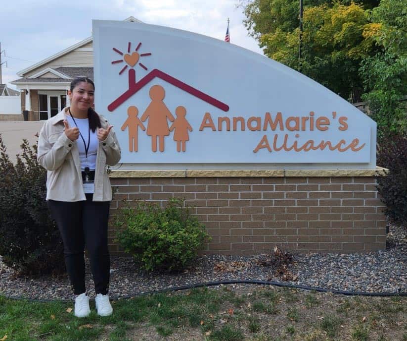 A woman stands smiling and giving two thumbs up in front of a sign that reads "AnnaMarie's Alliance." The sign features a stylized house with a heart and figures of an adult and children. The woman is dressed casually and stands on a gravel path with greenery around.
