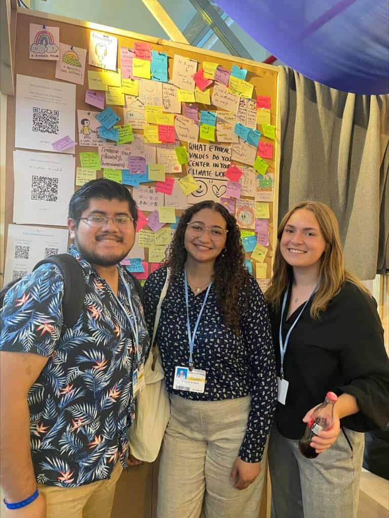 Three people stand smiling in front of a wall covered with colorful sticky notes and handwritten messages. The group looks cheerful and is dressed in casual to business-casual attire. One person holds a lanyard with an ID, and another holds a drink bottle.