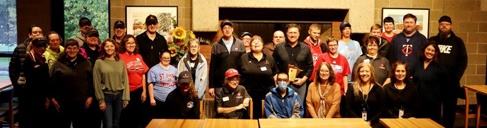 A group photo featuring numerous people standing and sitting together in what appears to be a community gathering or event. The group includes individuals of varied ages and attire, with some wearing name tags and uniforms. The setting includes tables and framed pictures.