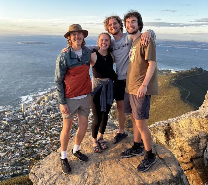 Four people are standing on a large rock overlooking a coastal city and ocean. They are smiling, and some have their arms around each other. One person is wearing a hat, another has a jacket around their waist. The sky is clear with scattered clouds.