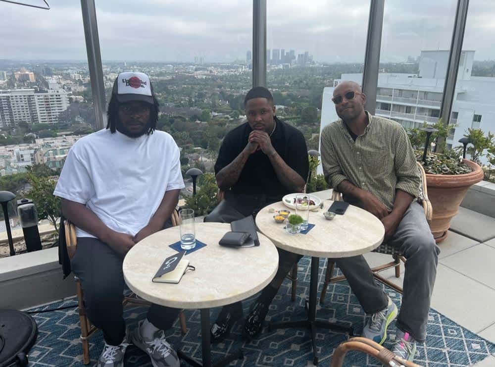 Three men sitting at a round table on a high-rise balcony with a cityscape in the background. The men, casually dressed, are engaged in conversation. The table has food, drinks, and phones on it. Large windows surround the balcony, offering a scenic view.