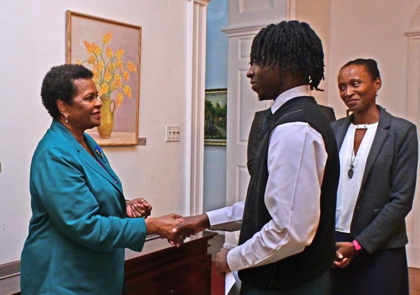 A woman in a teal blazer shakes hands with a man in a white shirt and black vest in a well-lit room. Another woman in a gray blazer stands nearby, smiling. A painting of yellow flowers hangs on the wall behind them.