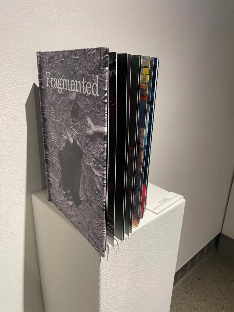 A book titled “Fragmented” with a grayscale textured cover stands open on a white pedestal. The exposed pages show vibrant colors and varied designs, suggesting mixed media artwork inside. The pedestal is placed against a plain white wall.