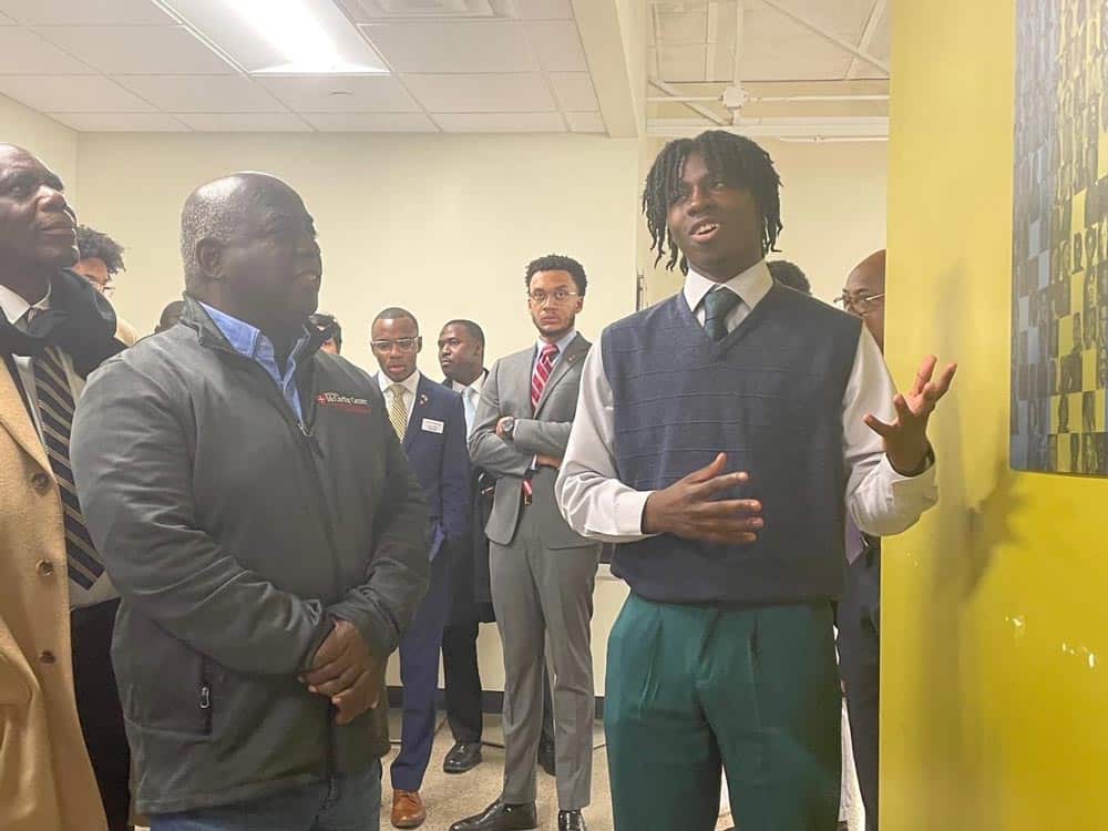 A young man with dreadlocks, wearing a sweater vest and green pants, passionately speaks to a group of attentive men, some in suits, gathered around him indoors. A painting is visible on the yellow wall to his right.