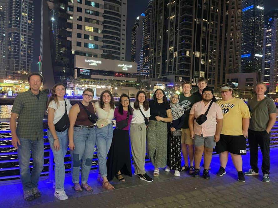 A group of thirteen people stand together smiling at night in front of a waterfront with illuminated buildings and skyscrapers in the background. Some are dressed casually, and others more formally. The scene is lively with bright city lights reflecting on the water.