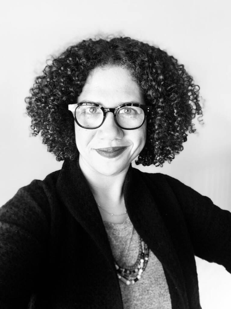 Black and white photo of a person with curly hair wearing glasses. They are smiling softly and wearing layered necklaces, a light-colored shirt, and a dark cardigan. They are facing the camera directly with a light background behind them.