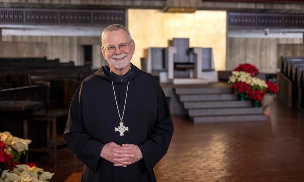 A smiling elderly man in clerical attire, wearing glasses and a large cross necklace, stands in a church with dark wood pews and red and white poinsettias near the altar. The background includes a raised platform with a high-backed chair.