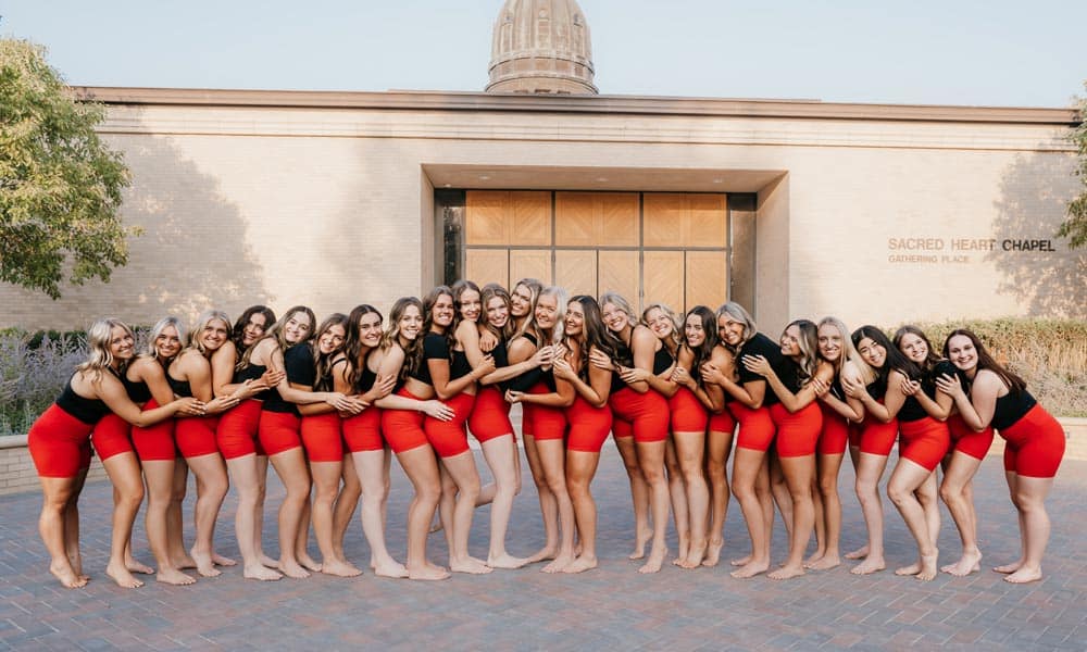A large group of women, dressed in black tops and red shorts, stand in a line in front of Sacred Heart Chapel. They are posing cheerfully with their arms around each other's shoulders. The background features a brick building and a dome structure.