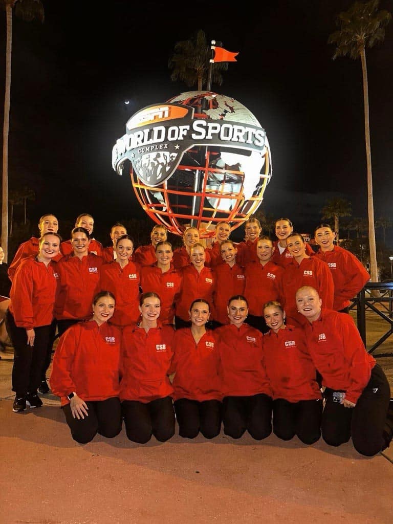 A group of people wearing matching red jackets poses for a photo in front of the ESPN Wide World of Sports Complex sign at night. The illuminated sign features a large globe with the complex's name on it, and palm trees are visible in the background.