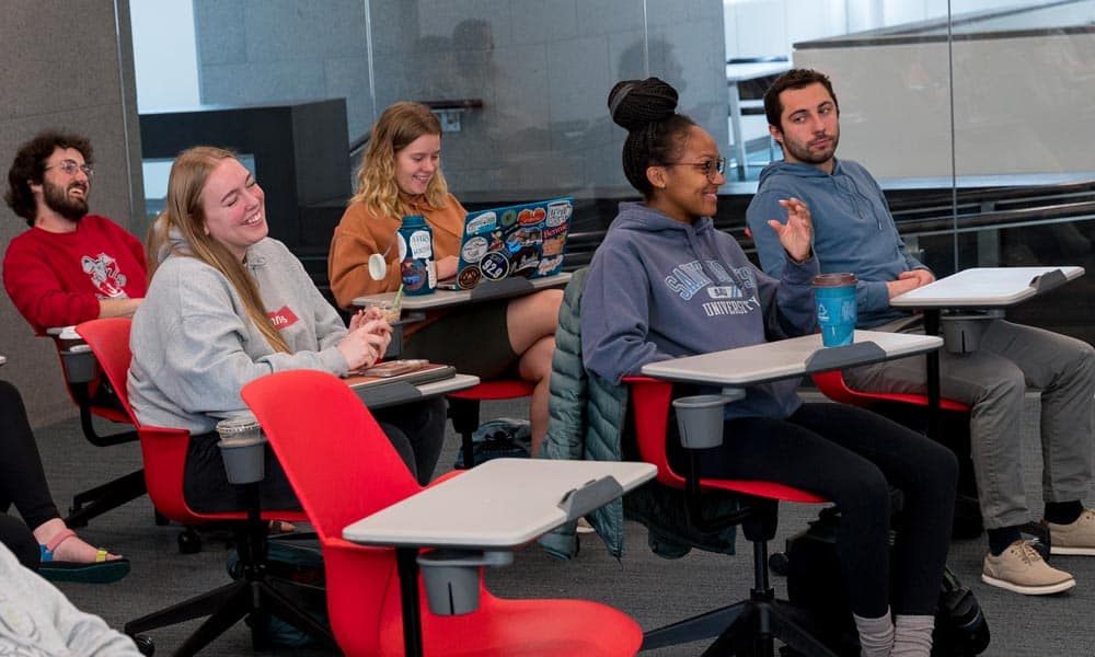 A diverse group of students sits in a modern classroom with red chairs and desks, attentively listening and engaging. One student, smiling, has a laptop covered in stickers. Another student raises their hand to speak, while others look on and participate enthusiastically.
