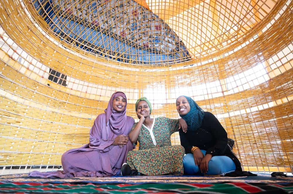 Three women sit closely together and smile inside a large, intricate wicker structure. The women are dressed in colorful, traditional attire, with one wearing a purple dress and hijab, the second in a green floral dress and hijab, and the third in a black dress and blue hijab.