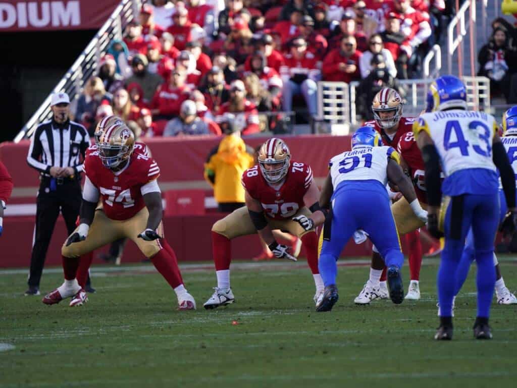 Football players in red uniforms prepare for a play against opposing players in blue uniforms on a green field. The stands are filled with spectators wearing red, and an official stands on the left. The focus is on the action at the line of scrimmage.