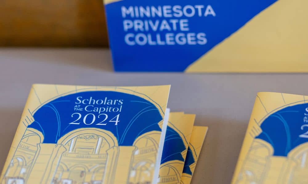 The image shows brochures titled "Scholars at the Capitol 2024" featuring an architectural line drawing of an interior space, likely a capitol building. In the background, there is a sign with a blue and yellow design that reads "Minnesota Private Colleges.