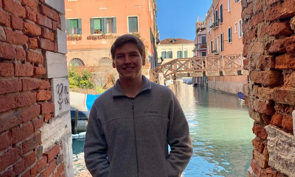 A man wearing a grey jacket stands smiling in a narrow alleyway. Behind him is a canal with clear blue water, bordered by red brick buildings and several boats. A wooden bridge crosses the canal in the background. The atmosphere is sunny and serene.