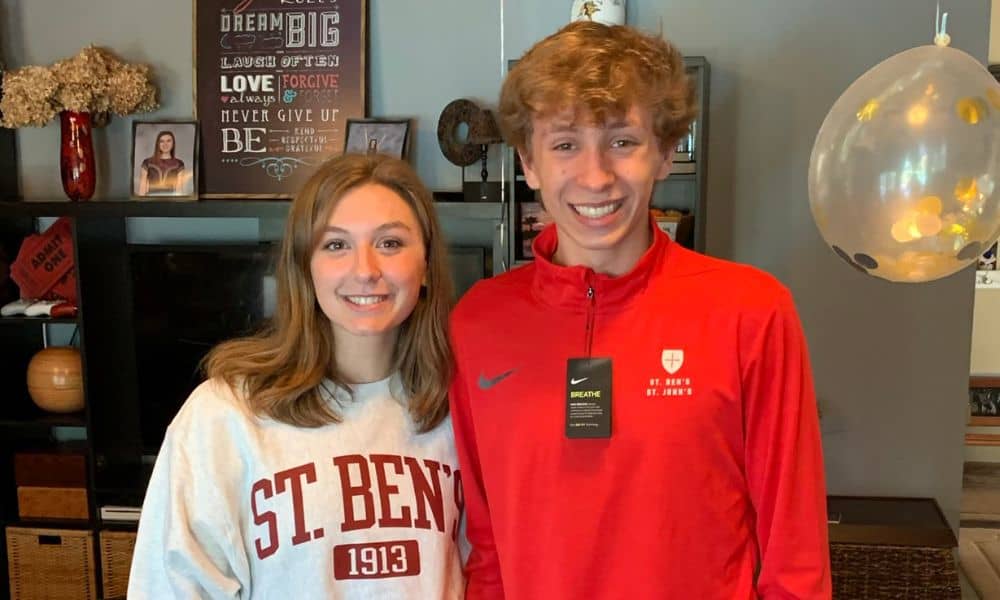 Two smiling individuals stand next to each other indoors. The person on the left is wearing a white "St. Ben's 1913" sweatshirt, while the person on the right is wearing a red "St. John's" jacket with a tag. They are in front of a shelf with decorations and a balloon.