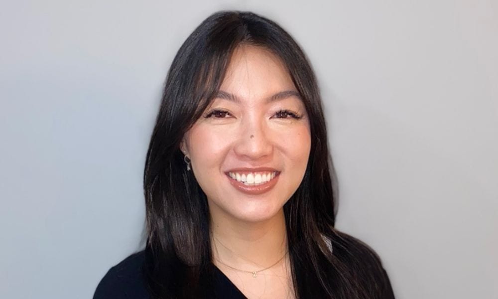A woman with long dark hair and a fringe is smiling at the camera. She is wearing a black top and a delicate gold necklace. The background is a plain light grey.