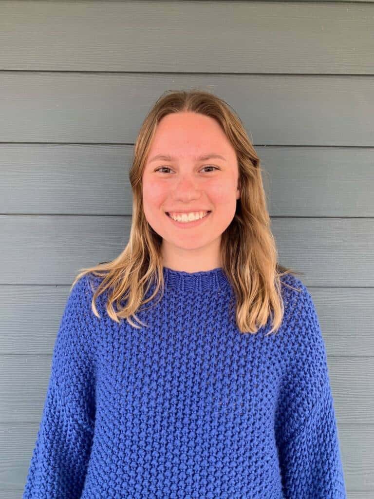 A person with shoulder-length blonde hair, wearing a bright blue knitted sweater, stands against a horizontally paneled light gray wall. They are smiling and looking directly at the camera.
