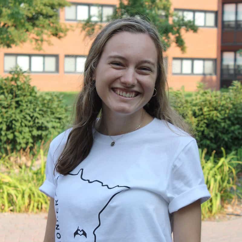 A smiling young woman with long hair wearing a white T-shirt and hoop earrings stands outdoors in front of a green bush with a building in the background. The T-shirt has a minimalistic design on it. She appears happy and relaxed.