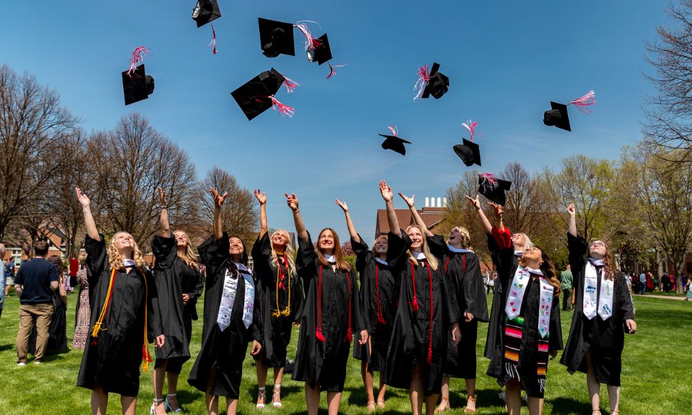 A group of graduates in caps and gowns celebrate by tossing their graduation caps into the air on a sunny day. They stand on a grassy area with trees and buildings in the background. Everyone is smiling and looking upwards at the caps.