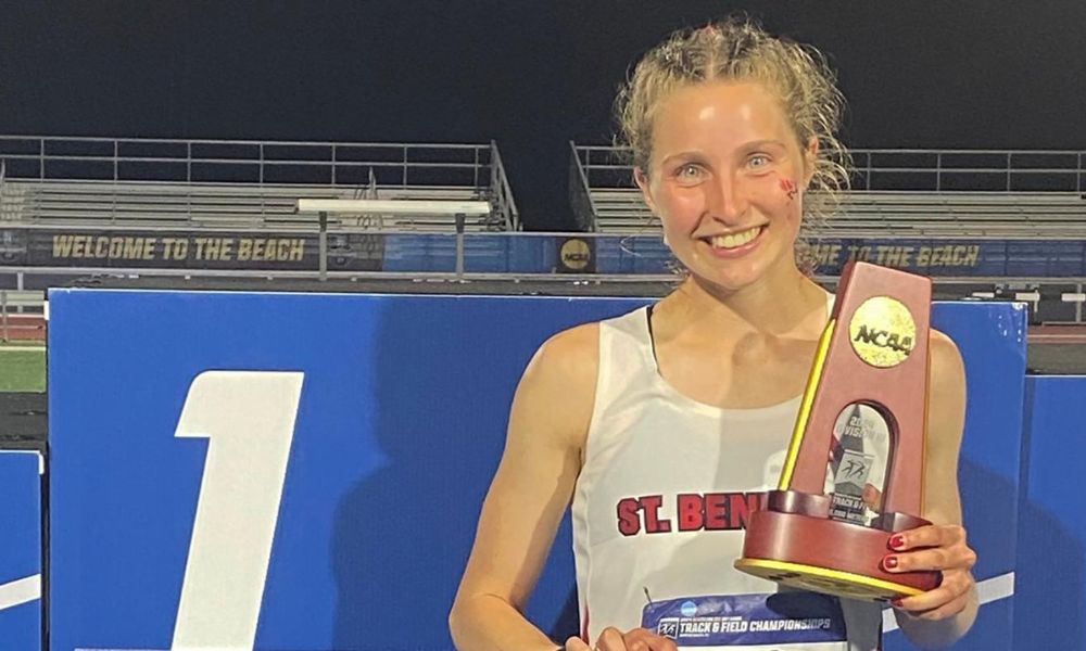 A young athlete wearing a "St. Benedictine" tank top smiles and holds an NCAA trophy in one hand and a rolled-up certificate in the other. She stands on a podium with the number 1, indicating her first-place victory. The background features stadium seating and a sign that says "Welcome to the Beach.