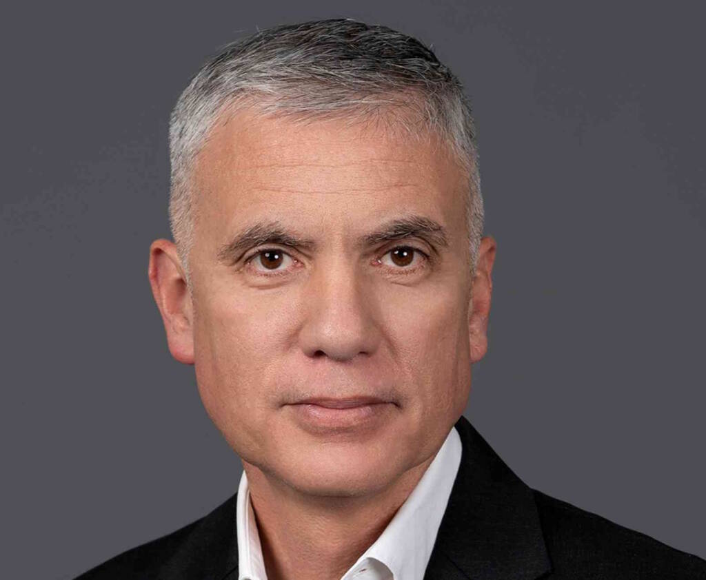 A man with short gray hair and brown eyes is wearing a dark suit jacket over a white dress shirt. He has a neutral expression and is posed against a plain gray background.