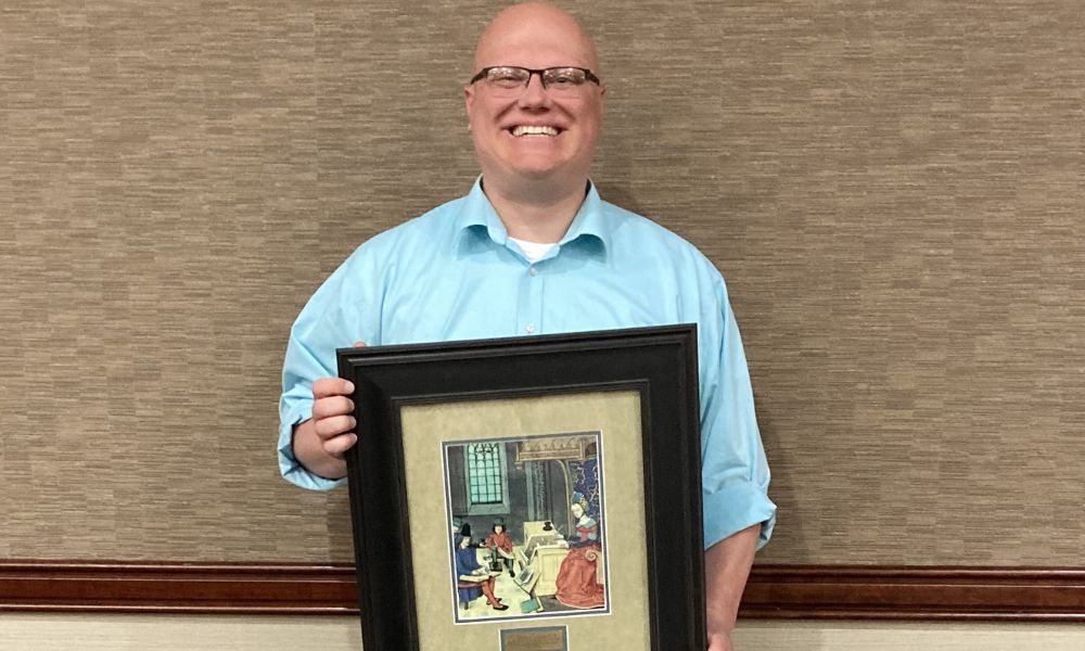 A smiling person with short hair and glasses stands against a beige wall, holding a large framed picture. The picture appears to feature an illustrated scene with several characters in a room. The person is wearing a light blue shirt.