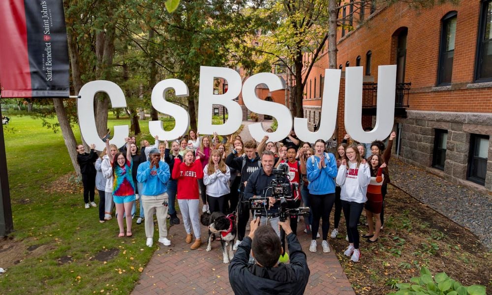 A large group of diverse college students stand outside, holding large letters forming "CSBSJU." They're smiling and cheering, with a camera crew filming them. The setting is on a campus with brick buildings and greenery in the background.