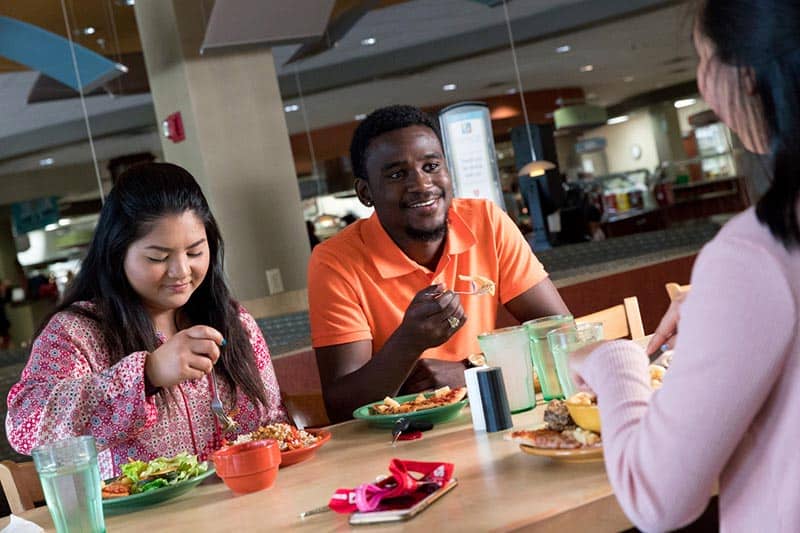 Three people are seated at a table in a cafeteria, enjoying a meal together. Two are facing the camera, smiling and eating, while the third person has their back to the camera. The table is set with plates of food and drinks. The background shows more cafeteria seating.