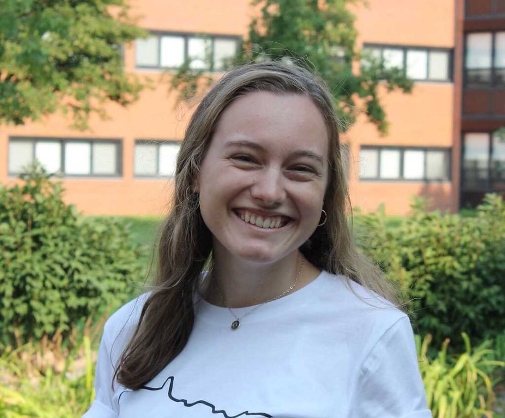 A young woman with long blonde hair is smiling broadly while standing outdoors. She is wearing a white shirt and a necklace. Greenery and a brick building with windows are visible in the background.
