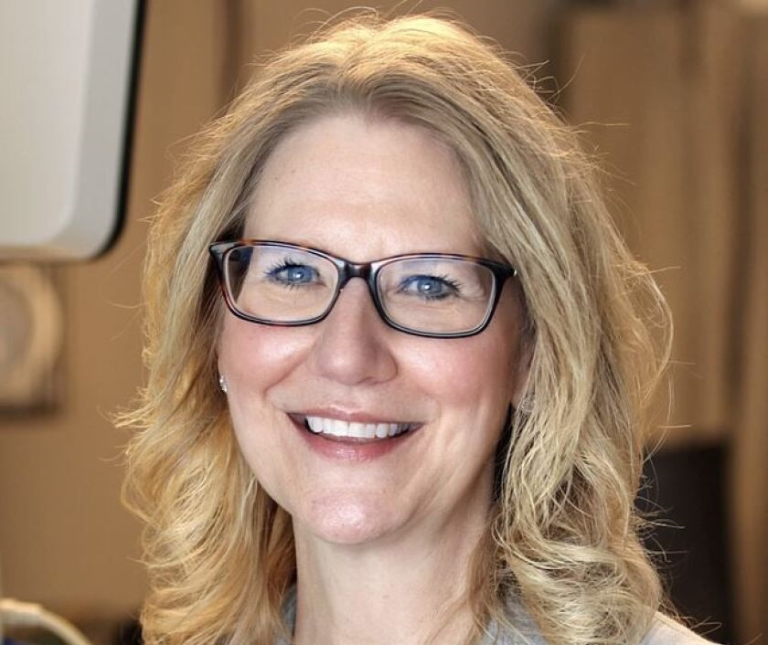 A woman with shoulder-length blonde hair and glasses is smiling. She is wearing a light-colored top and is photographed in a well-lit indoor setting.