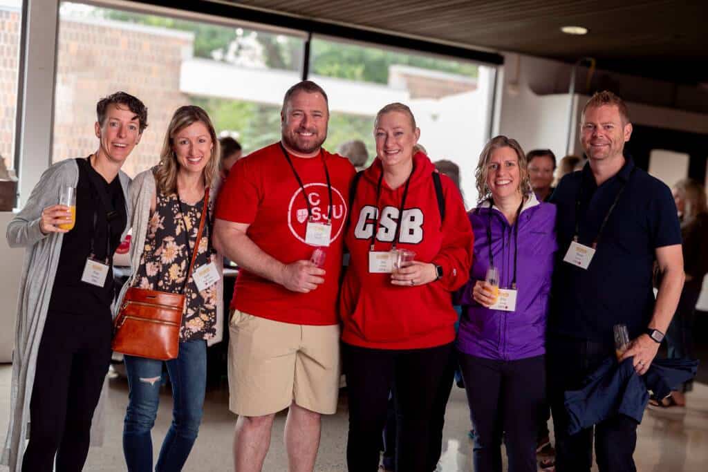 A group of six people pose for a photo indoors, smiling and holding drinks. They wear casual clothing and conference badges. Two people in the center wear red shirts, one with "CSU" and the other with "CSB" on it. The background features windows and blurred people.