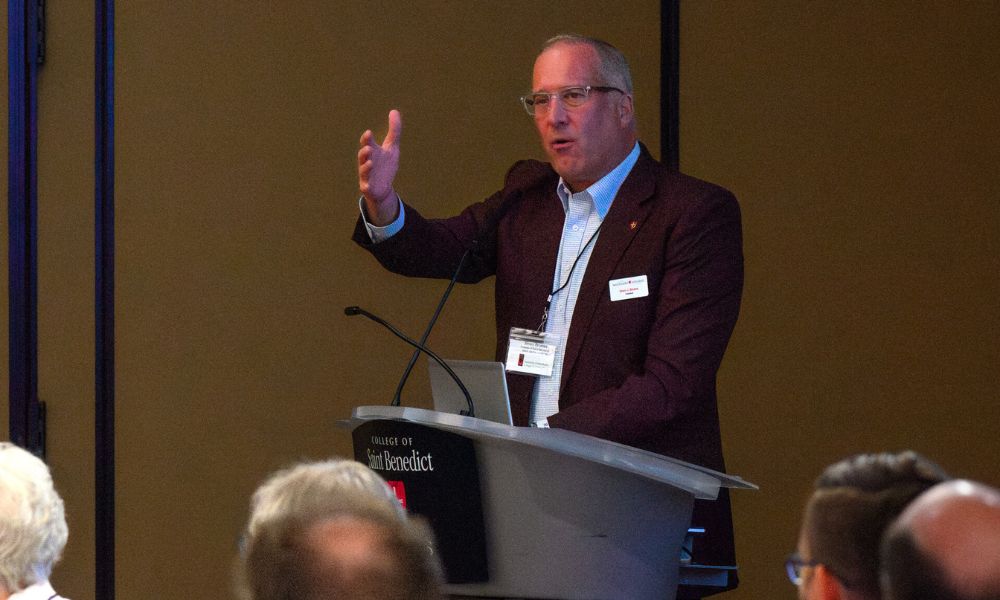 A man wearing glasses and a maroon jacket speaks at a podium, gesturing with his right hand. He has a name badge on his lapel. Several people are seated in the audience, and the background is a neutral-colored wall.