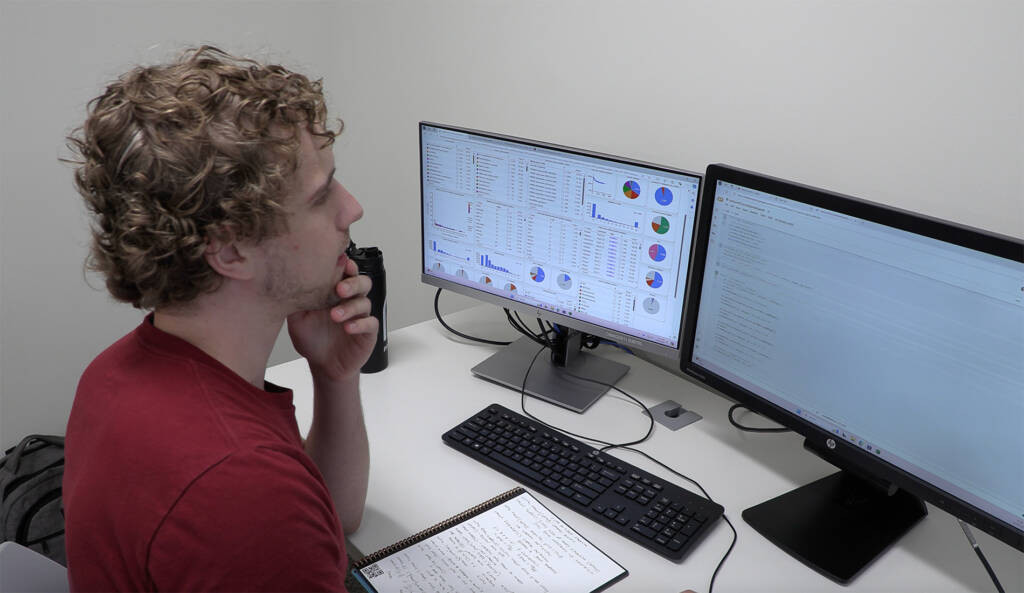 A person with curly hair, wearing a red shirt, sits at a desk with two computer monitors. One monitor displays various charts and graphs, while the other shows text documents. The person is looking at the screens and holding a pen, with a notebook open in front of them.