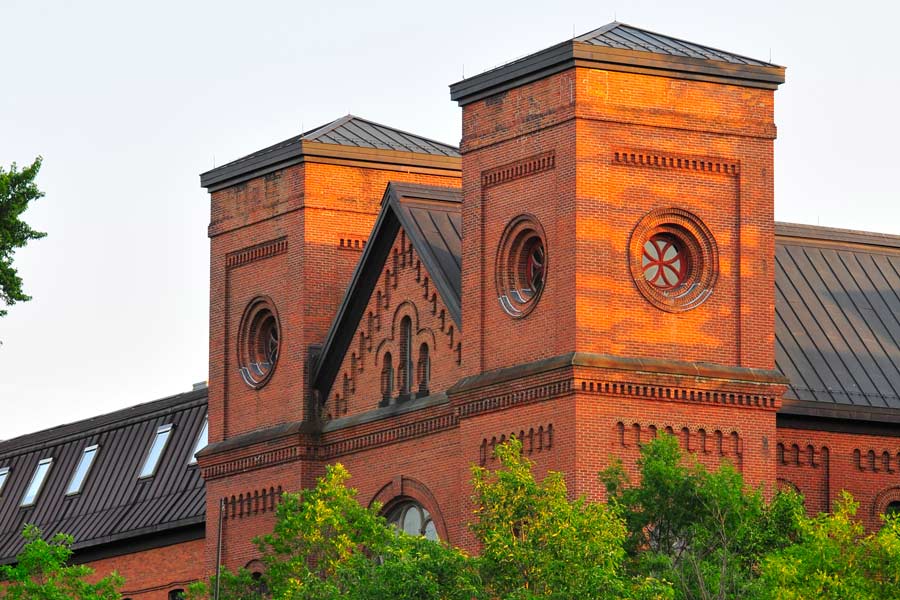 A photograph of a red-brick building with two prominent square towers. Each tower features a large circular window with decorative designs. The building has a steep, darkly colored roof and is partially obscured by green trees in the foreground.