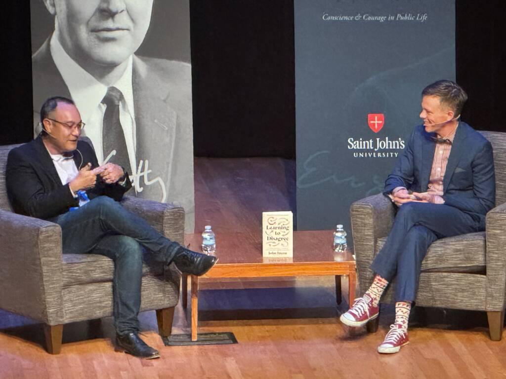 Two individuals are seated on stage having a conversation during a seminar at Saint John's University. A book is displayed on a table between them. Behind them is a large black and white image of a man and a Saint John's University banner.