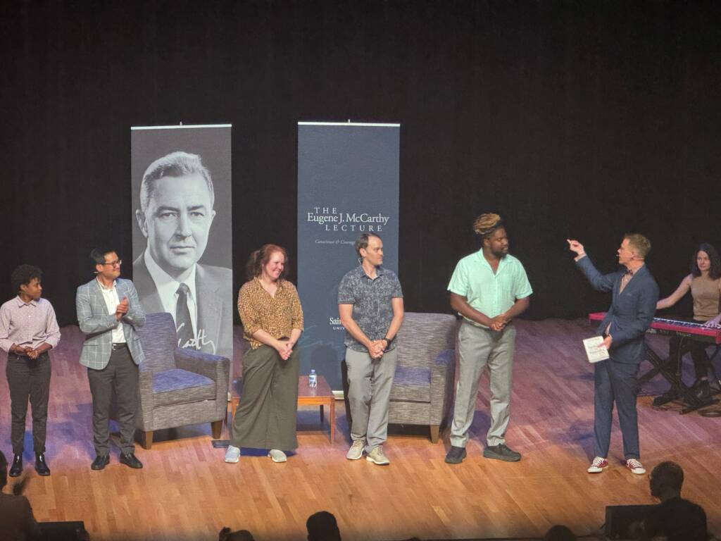 A group of five people stands on a stage next to two large posters and a screen displaying "The Eugene J. McCarthy Lecture." The moderator is engaging with the group while another person sits at a keyboard in the background. They are all in casual to semi-formal attire.