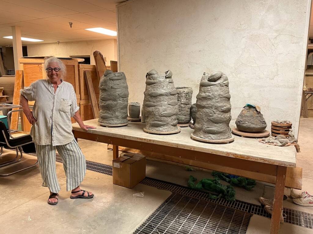 A person wearing striped pajamas and sandals stands smiling beside a table with several large, abstract clay sculptures in a workshop setting. The room has beige walls and various tools and materials in the background.