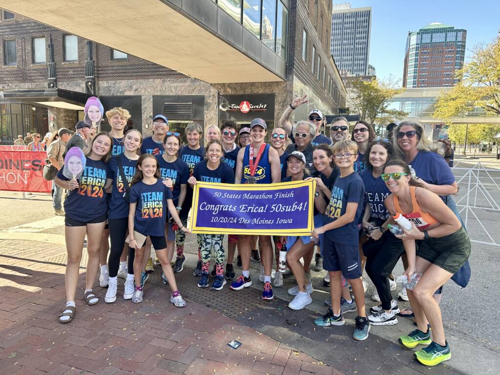 A group of people poses together outdoors, holding a congratulatory banner for a marathon finisher. Many are wearing matching "Team Erica" shirts. Buildings and trees are visible in the sunny background.