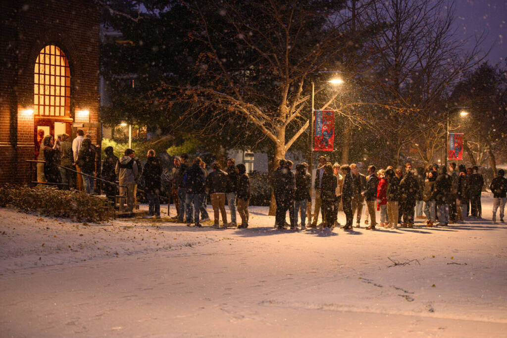 A group of people stand in line outside a building on a snowy evening. They are dressed in winter clothing, and streetlights illuminate the scene. Snow covers the ground and softly falls, creating a cozy, winter atmosphere.