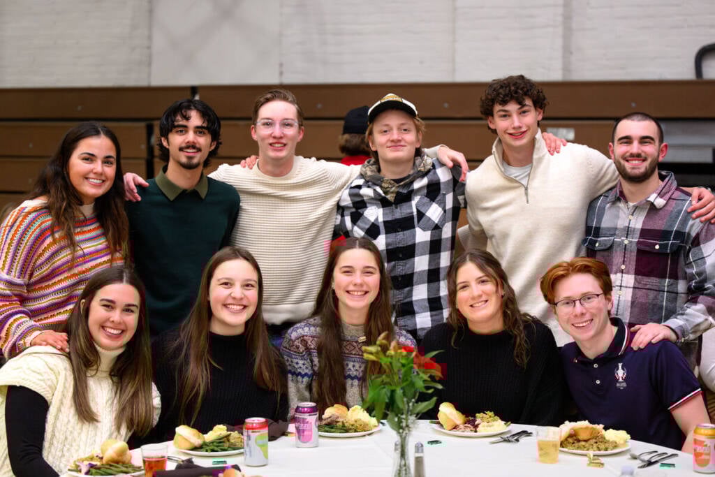 A group of people gathered around a table, smiling at the camera. Some are sitting, while others stand behind them. The table is set with plates of food, drinks, and a rose in a vase. They appear to be in a casual indoor setting.