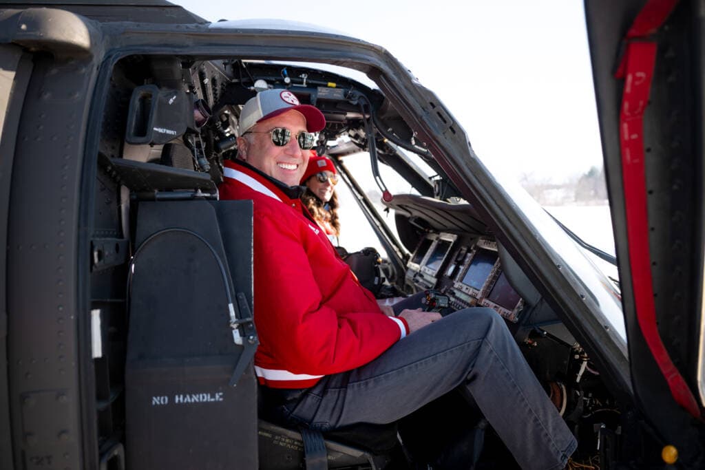 Two people smiling inside a helicopter cockpit. The person in the foreground is wearing a red jacket and sunglasses. The cockpit is detailed with various controls and displays. Snowy landscape visible through the windows.