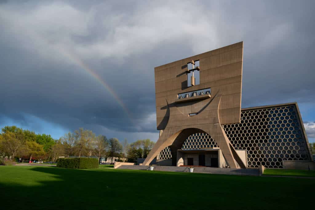 A modern architectural building with geometric patterns stands under a cloudy sky. A subtle rainbow arcs on the left, and the foreground features a well-maintained green lawn. Trees with newly sprouted leaves are visible in the background.