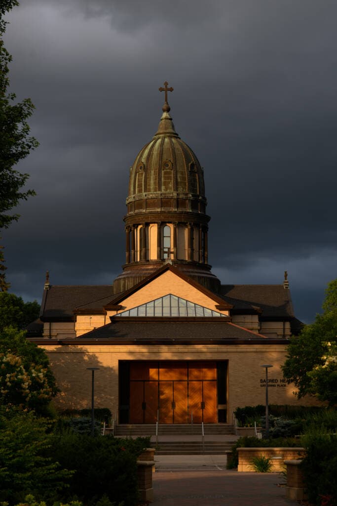A church with a tall, green-domed tower is bathed in golden light under a dark, cloudy sky. The entrance and facade are partially illuminated, contrasting with the stormy, dramatic background. Greenery surrounds the building.