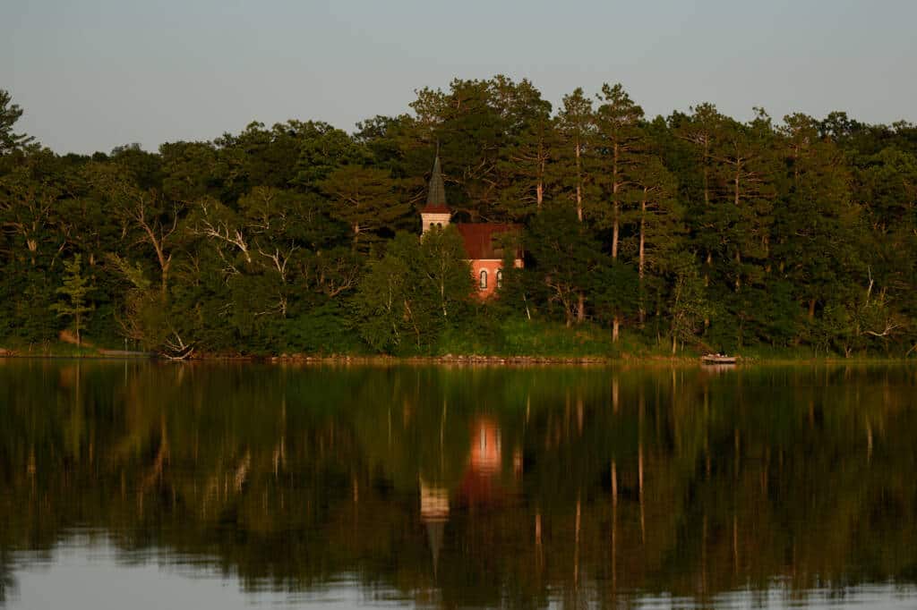 A small, red brick building with a spire is partially obscured by dense green trees on a lakeside. The calm lake reflects the trees and building in the golden light of sunset or sunrise.