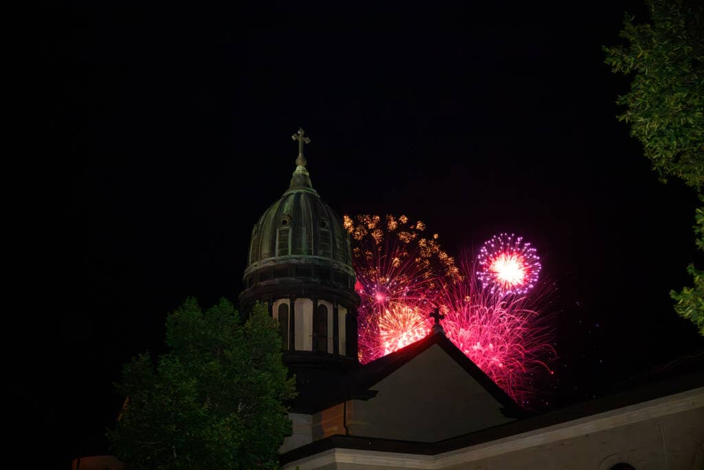 Fireworks burst behind a historic building with a domed roof and a cross on top. The night sky contrasts with the vibrant colors of the fireworks, and trees are partially visible in the foreground.