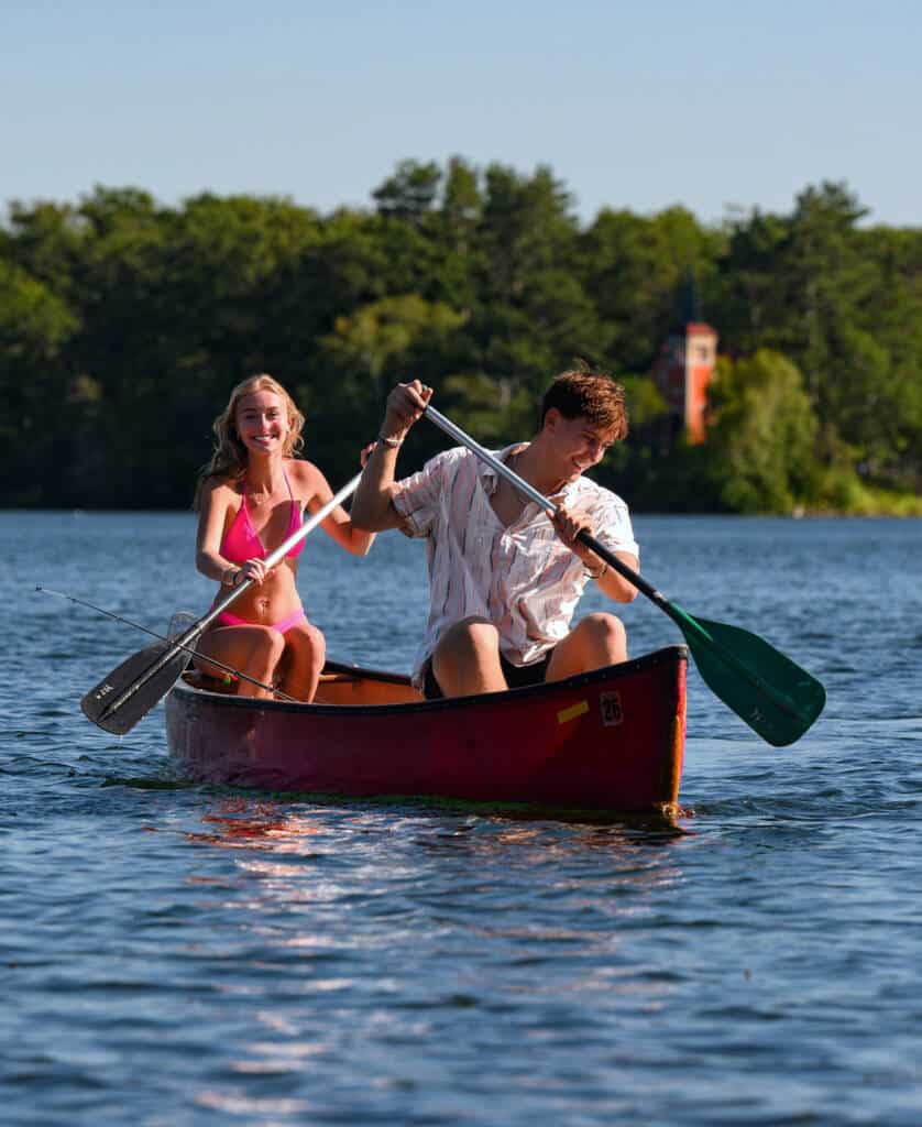 A young woman and man are joyfully paddling a red canoe on a calm lake, surrounded by lush green trees and a distant orange building. The sun is shining brightly, creating a lively summer atmosphere.