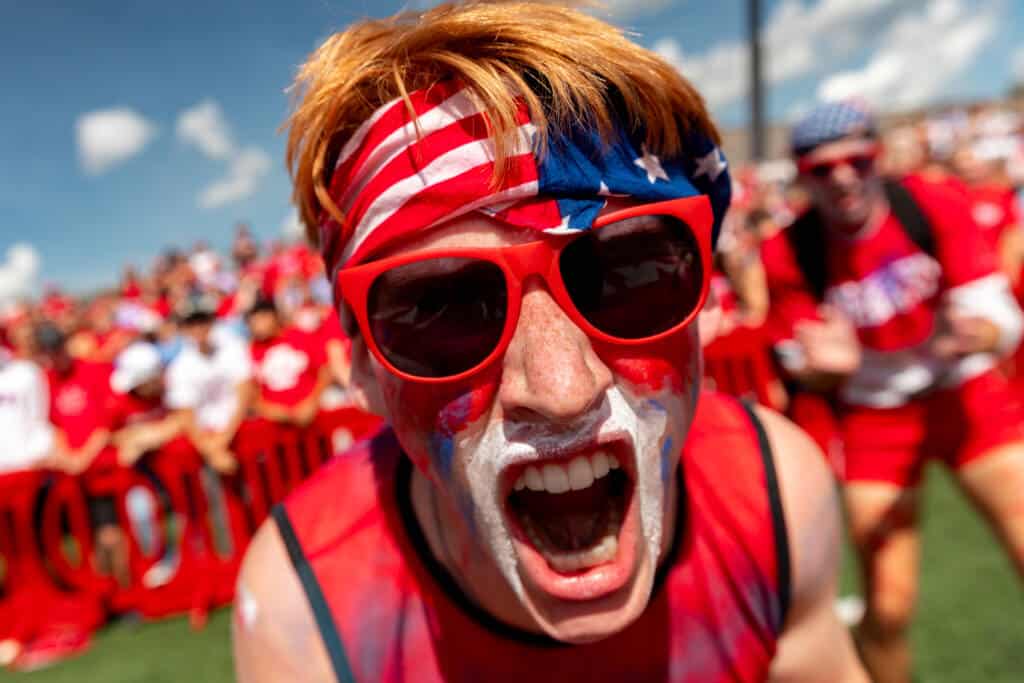 A person wearing red sunglasses, an American flag bandana, and sports-themed face paint passionately cheers in a crowd. The background shows more people dressed in red under a sunny sky.