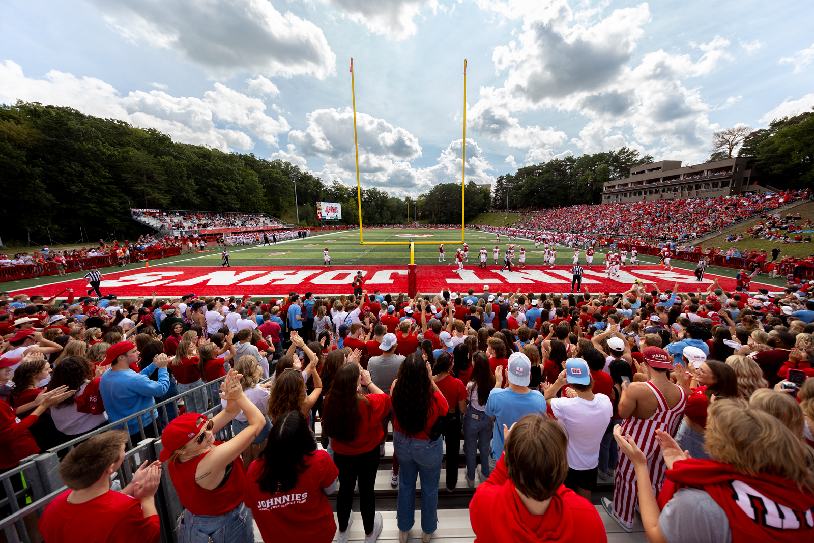 A large crowd of fans, mostly dressed in red and white, cheer at a college football game in a stadium. Players are on the field near the goalposts, with a bright red end zone labeled "Saint John's." The stands are filled under a partly cloudy sky.